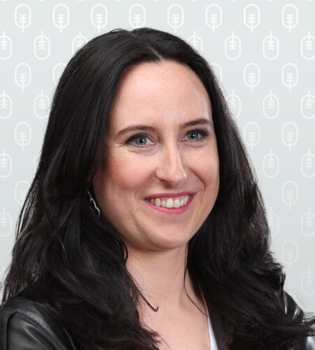 Eva steht mit verschränkten Armen und einer schwarzen Lederjacke mit neutralem Hintergrund da.